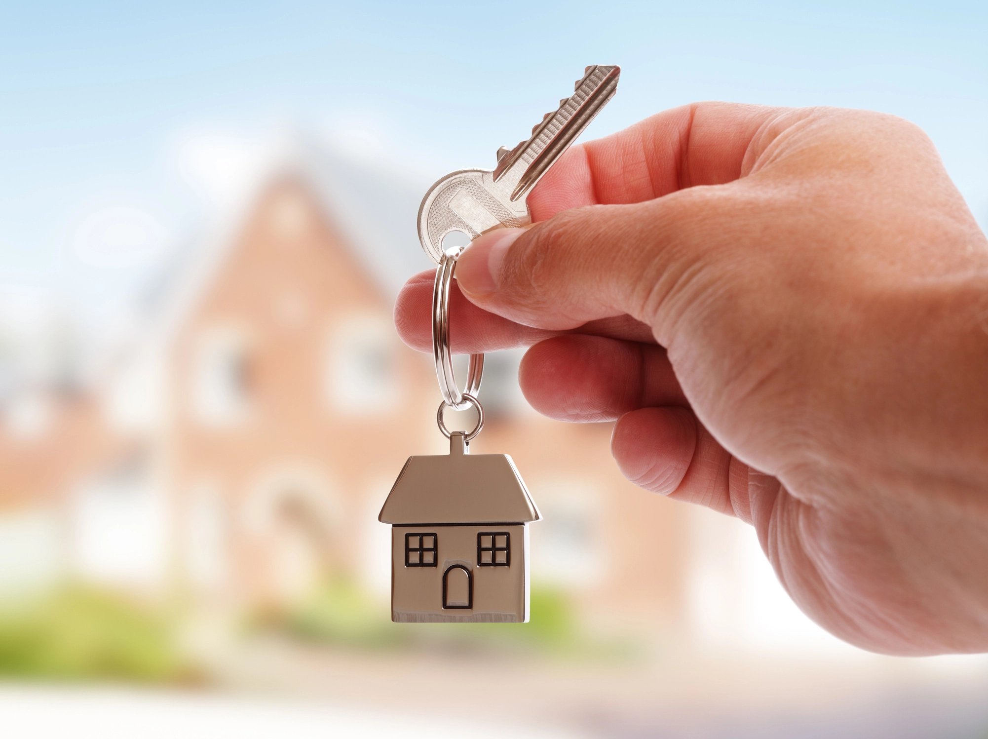 Hand holding house key with miniature house keychain, blurred home in background